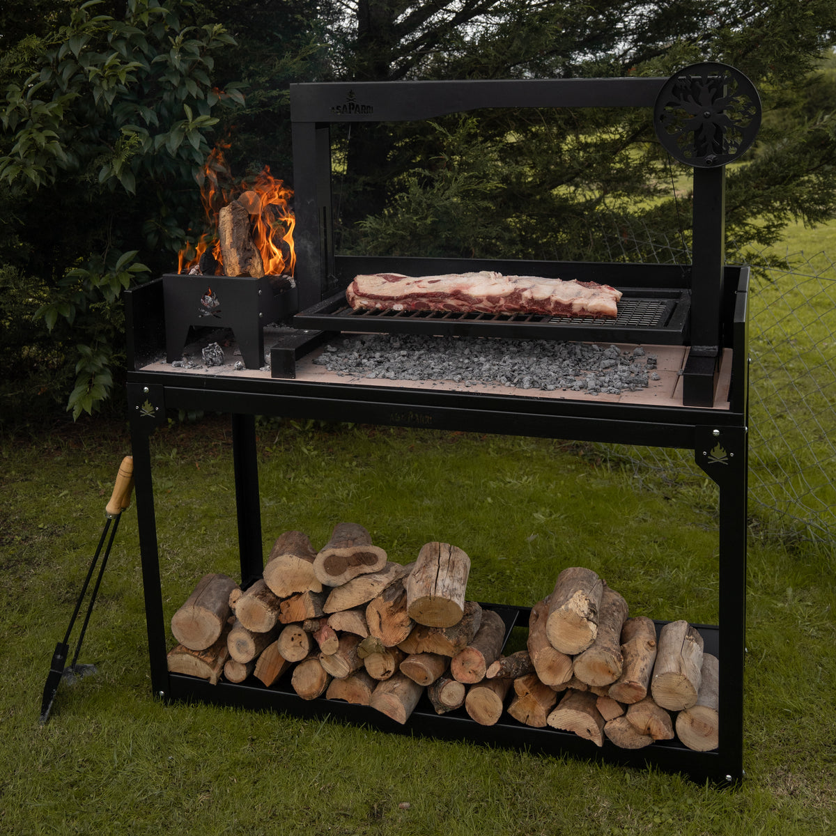 Meat cooking on the Asaparri Santa Maria grill outdoors, with a fire in the side brasero and logs below the base.