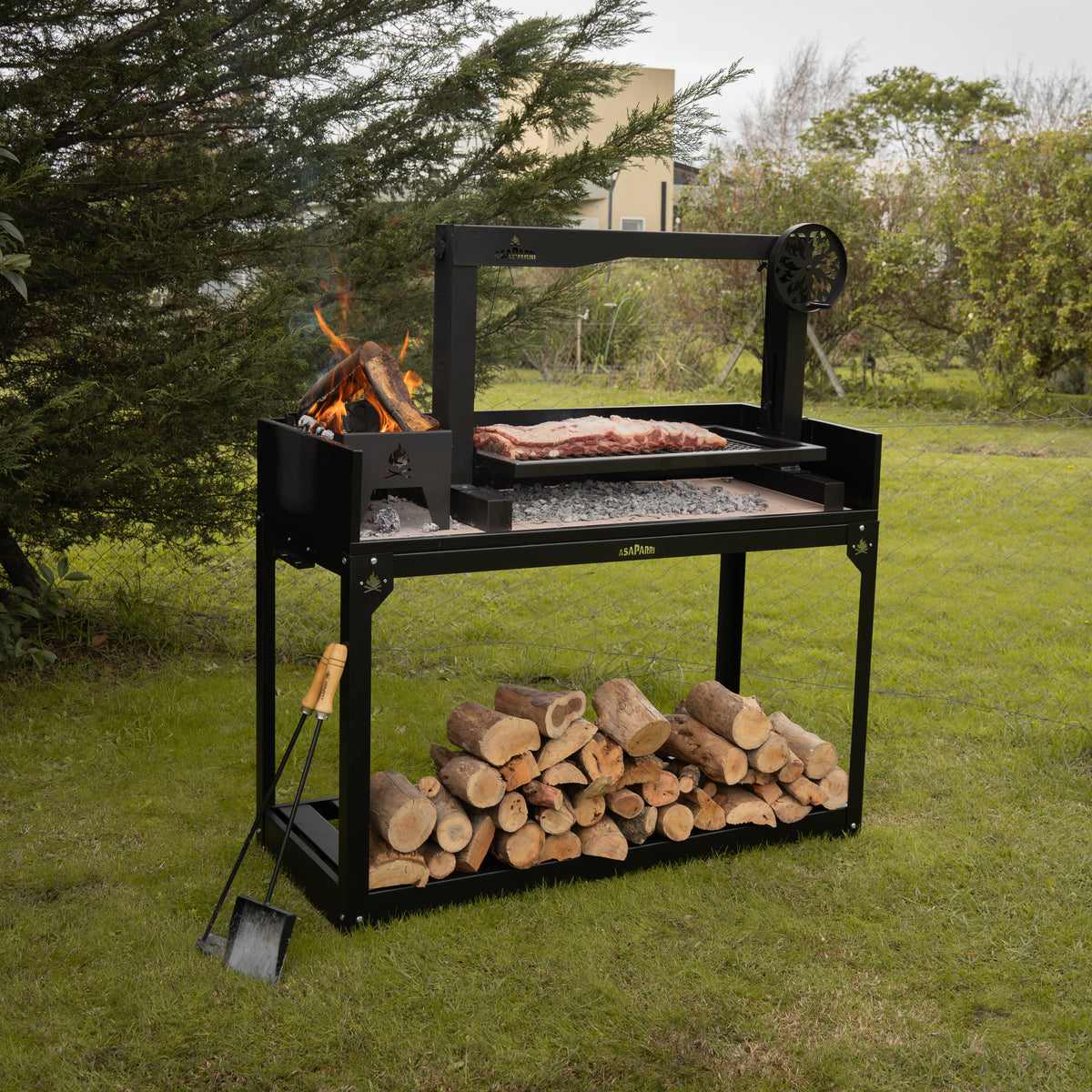 Asaparri Santa Maria Grill + Base on a lawn, with a fire burning in the brasero and logs stored underneath.